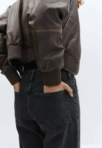 Chaqueta bomber de piel marrón, recortada, con puños acanalados, combinada con pantalones vaqueros de denim negros de cintura alta que tienen grandes bolsillos traseros.