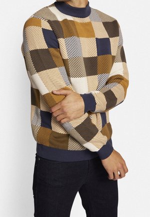 Homme portant un pull à carreaux multicolores avec des carrés bleu marine, moutarde, marron et beige, assorti à un pantalon foncé sur un fond uni.