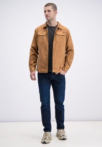 Hnědá semišová bunda se dvěma kapsami na prsou, v kombinaci s černou košilí, modrými džínami a béžovými teniskami, na jednobarevném pozadí.