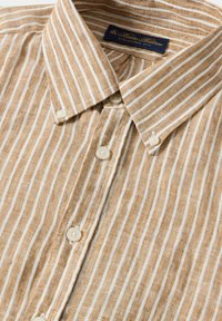 Camicia in lino a righe marroni e crema con colletto a bottoni, dotata di bottoni in plastica bianca e di un'etichetta blu all'interno del colletto.