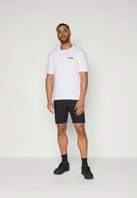 Balta kokvilnas t-kreklā ar īsām piedurknēm un mazu sarkanu logo, apvienots ar melniem šortiem un melnām sporta apaviem. Fons ir vienkāršs.