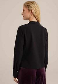 Blusa nera a maniche lunghe con taglio dritto, caratterizzata da polsini con bottoni e una texture liscia, abbinata a pantaloni in velluto viola.