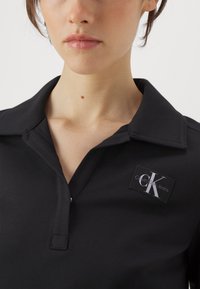 Fekete galléros póló, elülső gombolóval. Fehér Calvin Klein Jeans logó található egy téglalap alakú folton.