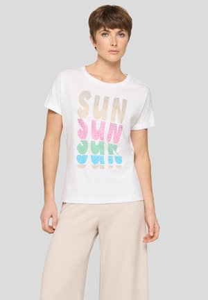 Frau mit kurzen braunen Haaren, die ein weißes T-Shirt mit buntem "SUN"-Schriftzug und beige Hosen mit weitem Bein trägt, steht vor einem grauen Hintergrund.
