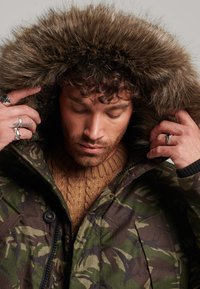 Parka de camuflaje con capucha forrada de piel; cuenta con cierre de cremallera y broches, tejido resistente y puños de canalé. Suéter de punto cálido y texturizado debajo.