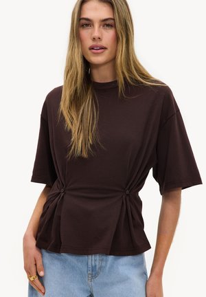 Mujer joven con cabello rubio largo que lleva una camiseta de manga corta marrón oscuro con botones laterales y vaqueros de mezclilla azul claro.