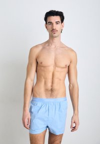 Svetloplavi bombažni bokserji z mehkimi pasom, na katerem je logotip Calvin Klein. Model ima fit postavo in stoji ob enobarvni ozadju.