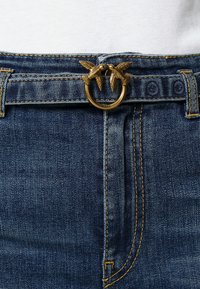 Primer plano de unos pantalones de mezclilla azules con un cinturón dorado que presenta dos hebillas en forma de pájaro detalladas en la cintura.