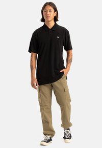 Polo nero con colletto classico e piccolo logo, abbinato a pantaloni cargo khaki con tasche laterali e sneakers nere.