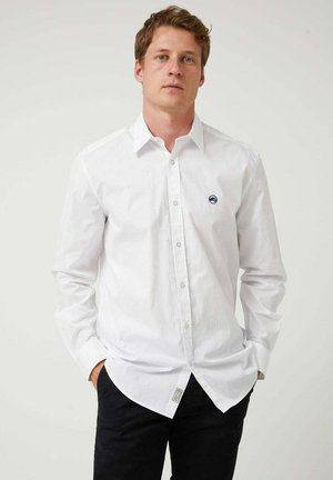 Joven con el pelo corto que lleva una camisa blanca abotonada con las mangas remangadas y pantalones negros, de pie con las manos en los bolsillos.