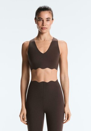 Kvinde iført en mørkebrun ærmeløs crop top og højtaljede leggings med takkede kanter, stående mod en ensfarvet hvid baggrund.