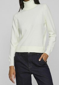 Pull à col roulé couleur crème en tissu doux, avec manches longues et coupe courte, associé à un jean en denim foncé.