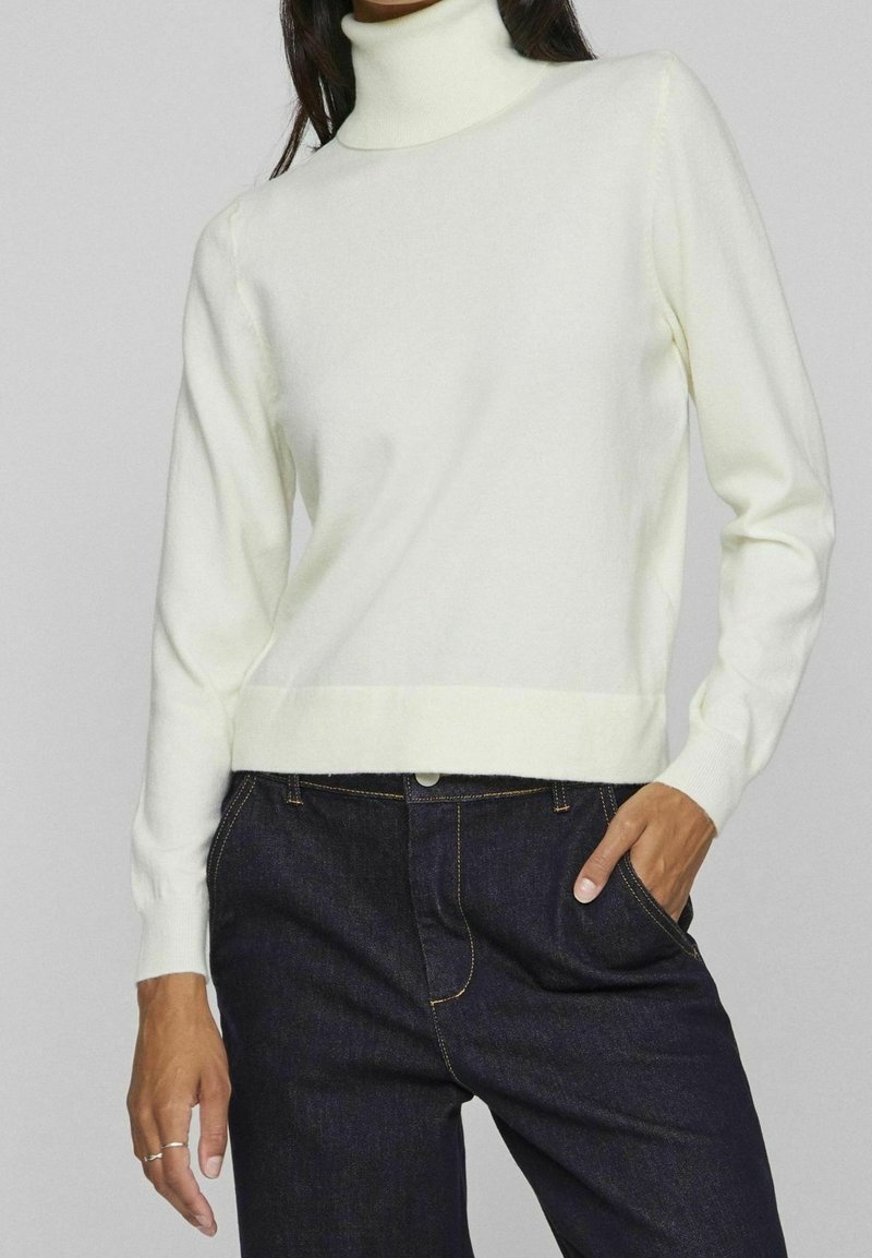 Pull à col roulé couleur crème en tissu doux, avec manches longues et coupe courte, associé à un jean en denim foncé.