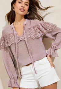 Light lavender blouse with lace trim and textured polka dots, featuring a V-neck and long sleeves with ruffled cuffs. Paired with white shorts.