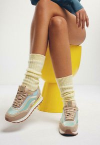 Zapatillas de deporte azul claro y beige con malla texturizada, suelas blancas y acentos amarillos, combinadas con calcetines de canalé color crema. Taburete amarillo al fondo.