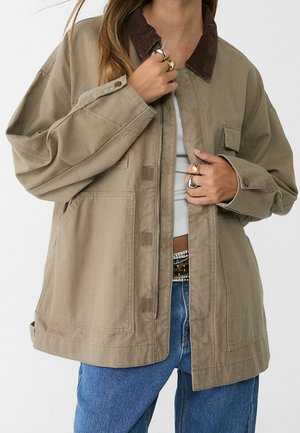 Person wearing a beige oversized jacket with a brown corduroy collar, blue jeans, and multiple rings on both hands.