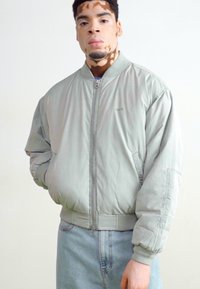 Gaiši pelēks bomber jaka ar rāvējslēdzēju priekšā, ribotām piedurknēm un apakšmalu; ar smalku logo uz krūtīm. Stilizēta ar gaišām džinsu biksēm.