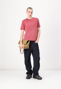 Rød gradient t-skjorte med trykt logo, mørkeblå jeans og en lys brune budveske. Svart treningssko med teksturdetaljer.