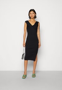 Vestido preto de malha canelada com decote em V, silhueta justa, fenda lateral e mangas tipo copinho. Usado com saltos verdes de tiras e uma bolsa estampada.