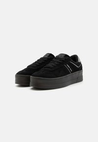 Baskets en suede noir avec un design plat, bout rond, lacets texturés et semelle en caoutchouc noir. Présente un logo sur le côté et la languette.