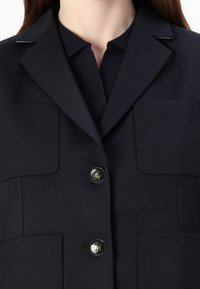 Granatowa marynarka z dwoma przednimi kieszeniami, klapami notch i guzikami w kolorze złotym z monogramem. Materiał ma gładką teksturę.