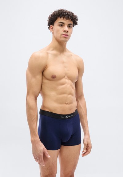 Νεαρός άνδρας με τα κυματιστά μαλλιά, φορώντας μποξεράκια Ralph Lauren σε ναυτικό μπλε χρώμα, στέκεται μπροστά σε απλό ανοιχτό γκρι φόντο, κοιτώντας ελαφρώς προς τα δεξιά.