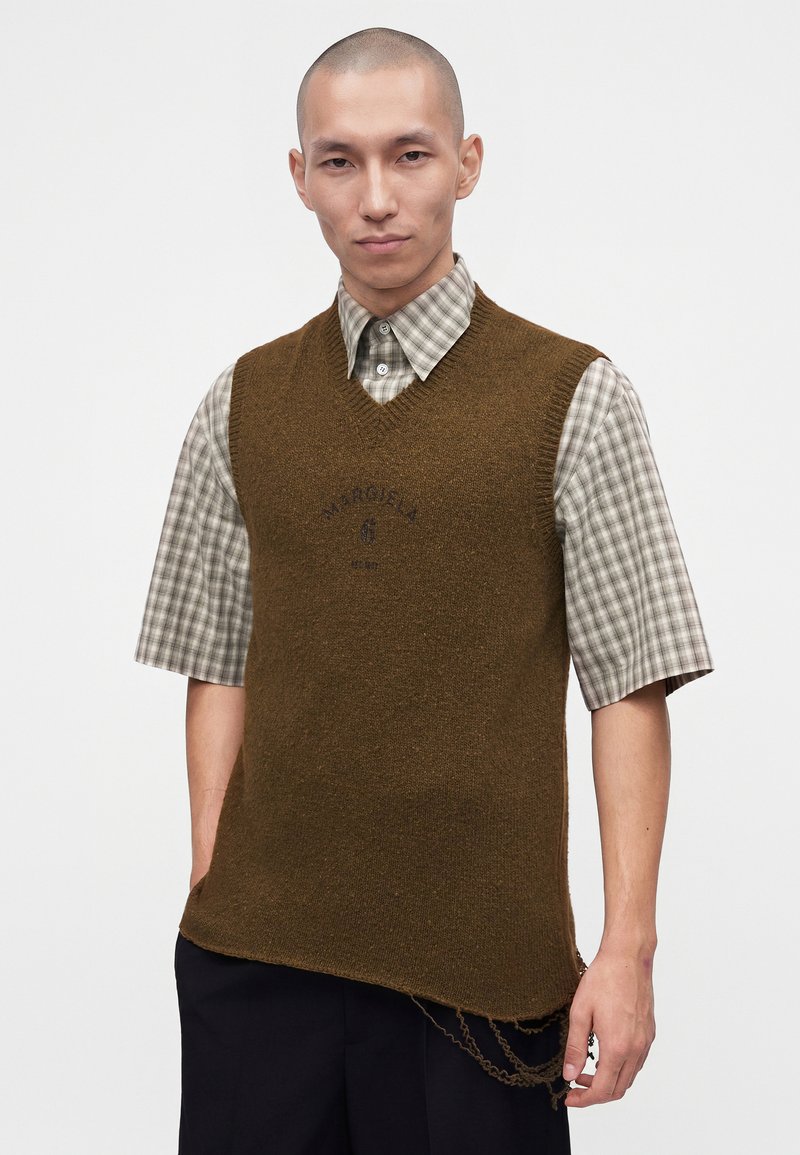 Mann trägt eine braune ärmellose Strickweste über einem taupe-weiß karierten kurzärmligen Hemd, mit schwarzen Hosen vor einfachem Hintergrund.