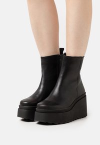 Bottines en cuir noir avec une semelle épaisse en coin, bout rond et fermeture éclair sur le côté pour un enfilage facile. Fini en cuir texturé avec des accents mats.