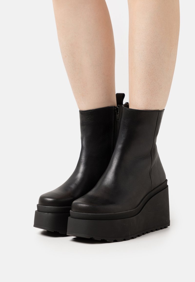 Bottines en cuir noir avec une semelle épaisse en coin, bout rond et fermeture éclair sur le côté pour un enfilage facile. Fini en cuir texturé avec des accents mats.