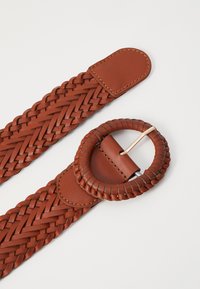 Woven leather belt in warm brown, featuring a circular belt loop and a smooth, rectangular strap end. Textured surface with braided details.