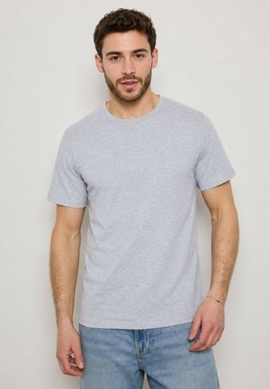 Homme aux cheveux courts, bouclés et foncés avec une barbe, portant un t-shirt gris clair à manches courtes et un jean bleu clair, debout devant un fond uni.