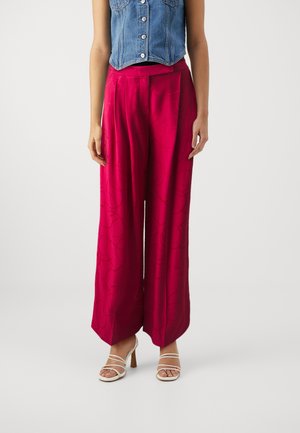 Femme portant un pantalon large taille haute magenta à motif subtil, des talons blancs à brides et un gilet en denim court sans manches.