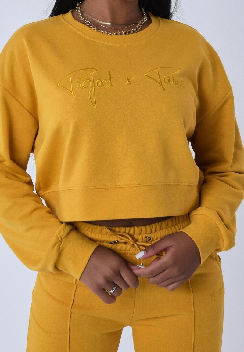 Femme portant un sweat court jaune moutarde et un pantalon de survêtement assorti, avec un collier chaîne en or et des bagues aux doigts.