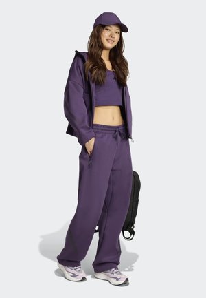 Conjunto atlético púrpura que incluye un top corto, chaqueta con cremallera y pantalones sueltos. Acentuado con una gorra púrpura y zapatillas deportivas grises.