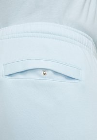 Pantalones de chándal azul claro con un bolsillo trasero con solapa, cerrado por un botón de presión. La tela parece suave y lisa, con un corte holgado.