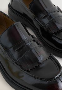 Black patent leather loafers with fringe detail, rounded toe, and stitched accents. Smooth texture and sleek design. Rubber outsole.