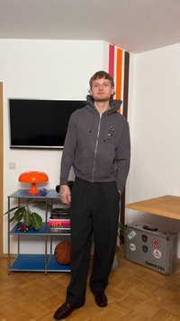 Ung mand iført en grå hættetrøje og sorte bukser står i et rum med et TV, en orange lampe, en bogreol, en basketball og en kuffert mærket Tropicana.