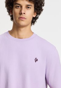 Levandų spalvos medvilninė marškinė su apvalia iškirpte ir trumpomis rankovėmis, puošia mažas tamsus siuvinėjimas kairiajame krūtinės kampe.