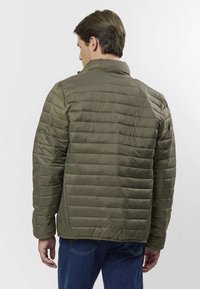 Geacă matlasată kaki, cu guler înalt, manșete drepte tricotate, tiv drept și textură netedă. Purtați cu blugi bleu închis.