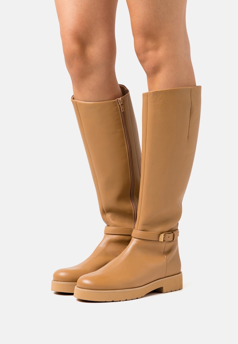 Bottes hauteur genou couleur camel en cuir lisse, dotées d'une fermeture éclair latérale, d'une sangle étroite avec une boucle métallique, et d'une semelle plate texturée.