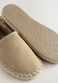 Zapatos slip-on de ante beige con suela de goma texturizada y borde de yute trenzado, que muestran un diseño simple y liso.