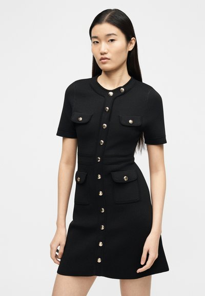 Γυναίκα που φοράει μαύρο φόρεμα με κοντά μανίκια, κλείνει με κουμπιά, έχει τέσσερις τσέπες με καπάκια και μεταλλικά κουμπιά, στέκεται μπροστά σε απλό φόντο.