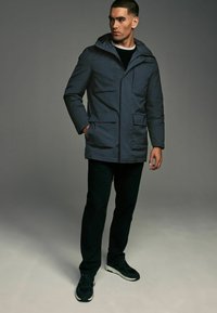 Parka acolchada de color azul oscuro con capucha, cuenta con múltiples bolsillos frontales y cierre de cremallera. Combinada con pantalones negros y zapatillas.