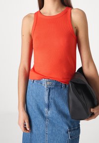 Top sin mangas de color rojo con textura acanalada y escote redondeado, combinado con una falda de mezclilla y un bolso de mano negro. La superficie tiene un aspecto suave y ajustado.