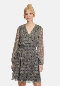 Schwarz-weiß gemustertes Kleid mit V-Ausschnitt, langen transparenten Ärmeln und Gummizug an der Taille, das einen ausgestellten Rock und geometrische Designs aufweist.