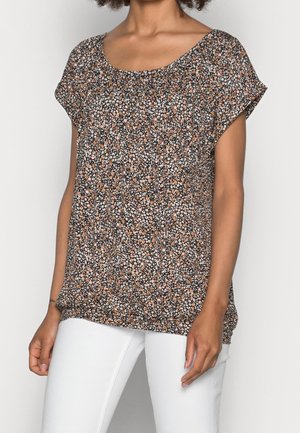 Femme portant un haut noir ample à motifs floraux avec de petites fleurs blanches et orange, associé à un pantalon blanc, se tenant devant un fond uni.