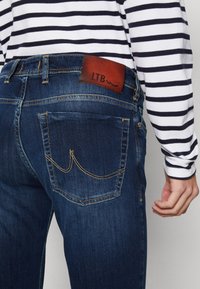 Vaqueros de mezclilla en azul oscuro con costuras amarillas. Presentan una etiqueta de cuero con el logotipo de LTB y bolsillos traseros con una costura decorativa.