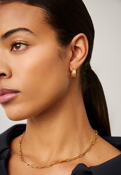 Γυναίκα με χρυσά σκουλαρίκια τριπλού κύκλου και χρυσή αλυσίδα στο λαιμό, με σκούρα μαλλιά που είναι τραβηγμένα προς τα πίσω, φοράει μαύρη μπλούζα.