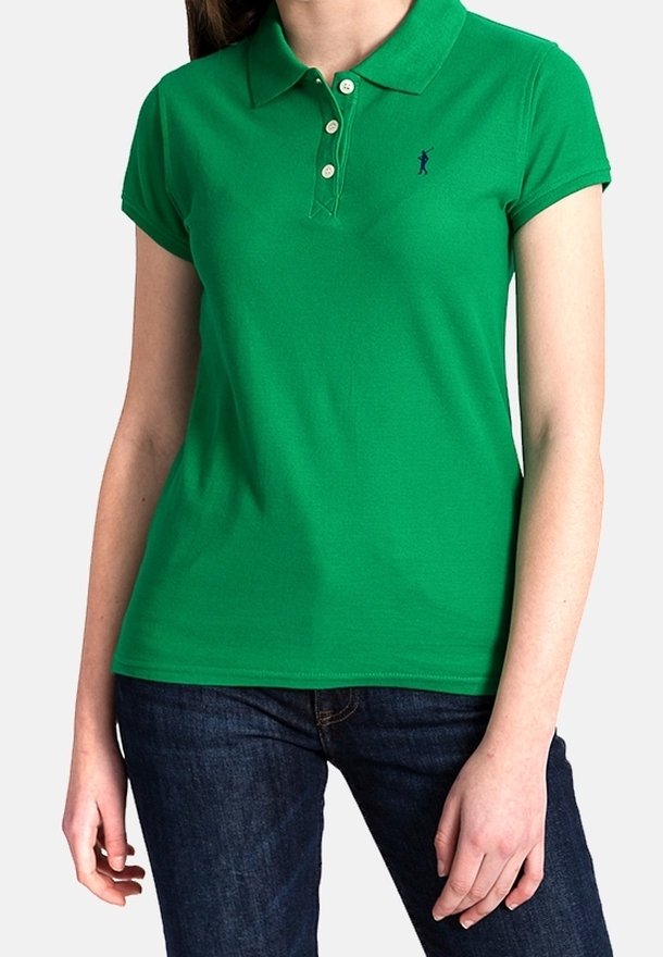 Femme portant un polo vert ajusté à manches courtes avec trois boutons et un jean bleu foncé, debout devant un fond uni.