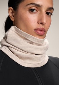 Bézs színű, puha anyagból készült nyakmelegítő, sima textúrával és réteges kialakítással; fekete sportfelsővel viselve, amely cipzárral rendelkezik.